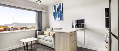 A modern living room with a sofa, a small table, a kitchen area with a counter, and a large window offering a view of the city.