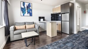 A modern living room with a grey sofa, a small table, and a kitchen area with a fridge and cabinets.