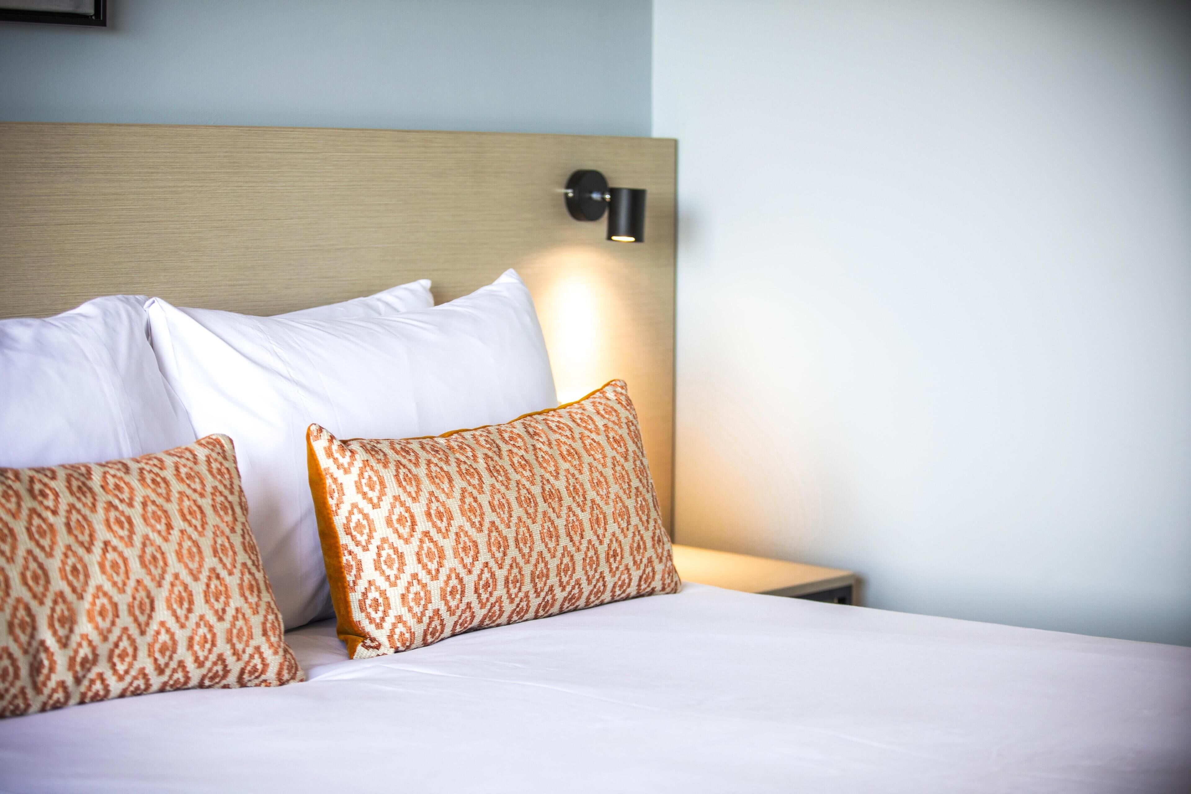A neatly made bed with white and patterned pillows, a wooden headboard, and a wall-mounted light fixture.