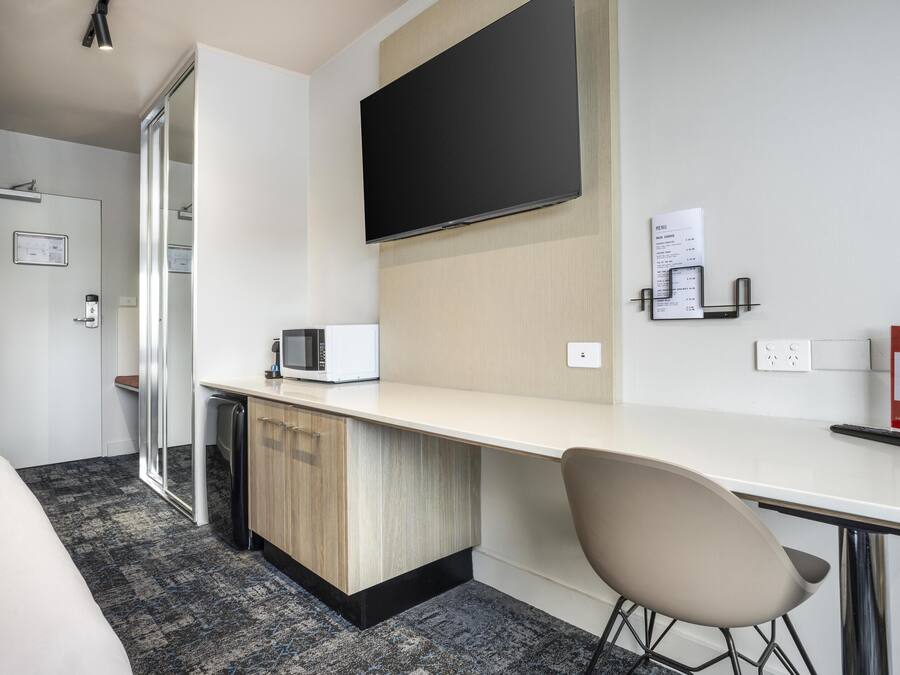 A hotel room with a bed, a desk with a chair, a microwave, a flat-screen TV, a door with a glass panel, and a wall-mounted menu board.