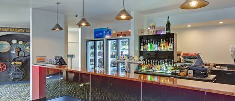 A bar area with a counter, overhead lighting, a refrigerated display, and a shelf with bottles.