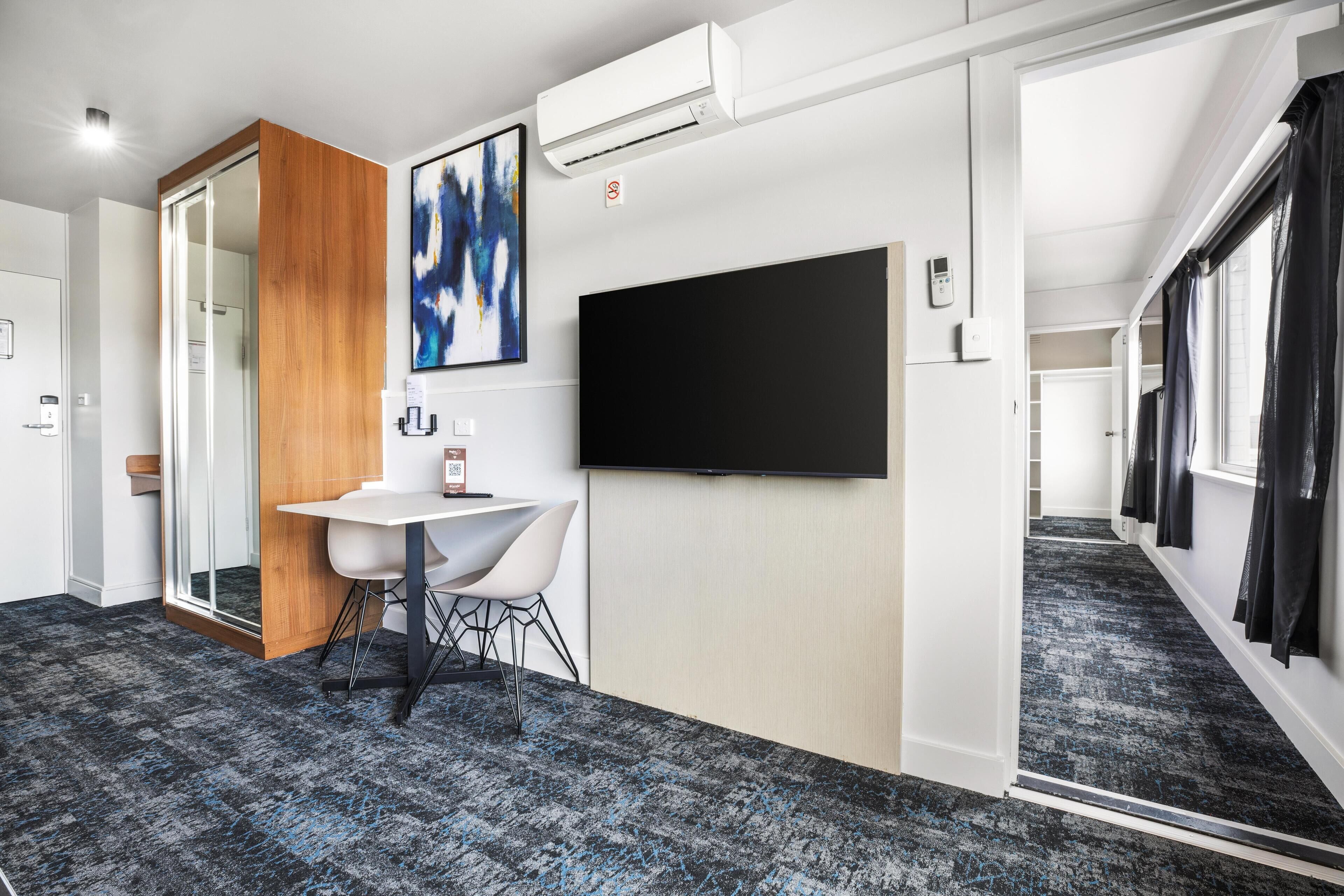 Una habitación de hotel moderna con un televisor de pantalla plana grande, una pared decorativa de madera, un escritorio blanco con silla y piso cubierto de alfombra.