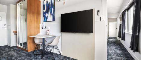 A modern hotel room with a large flat-screen TV, a wooden accent wall, a white desk with a chair, and a carpeted floor.