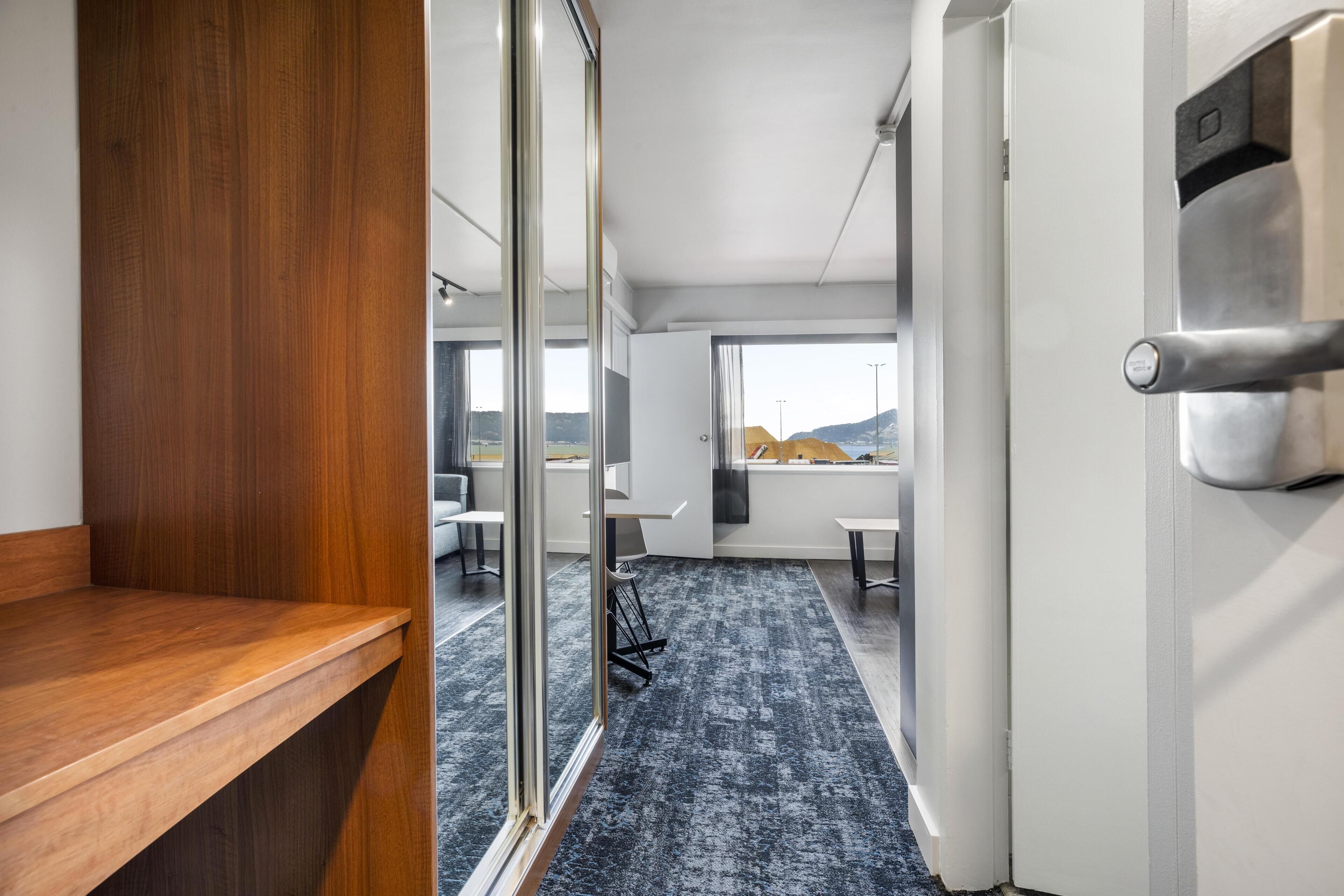 A modern hotel room with a wooden desk, a glass partition, and a view of the outdoors.
