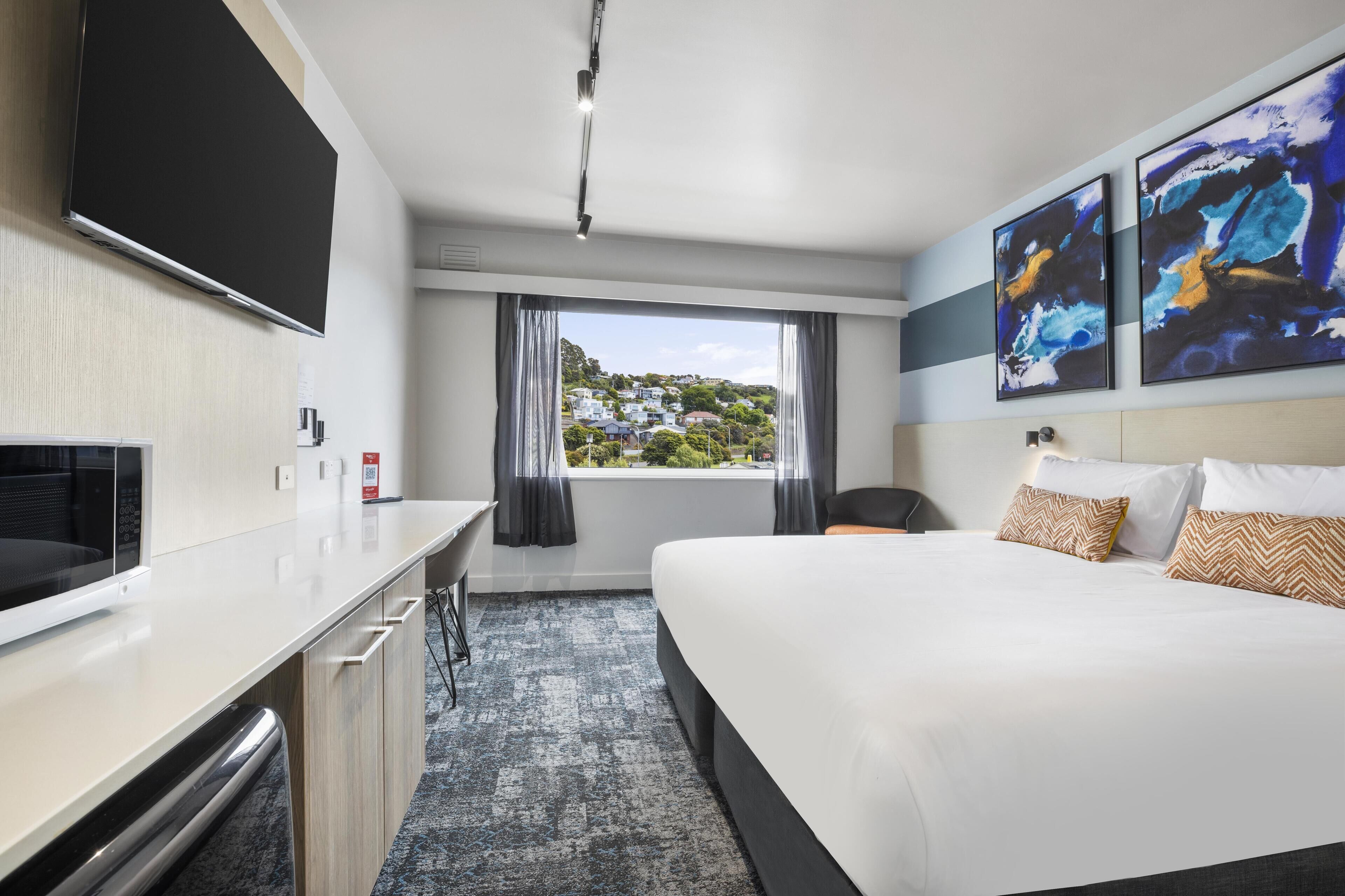 A hotel room with a bed, a desk, a flat-screen TV, and artwork on the wall.