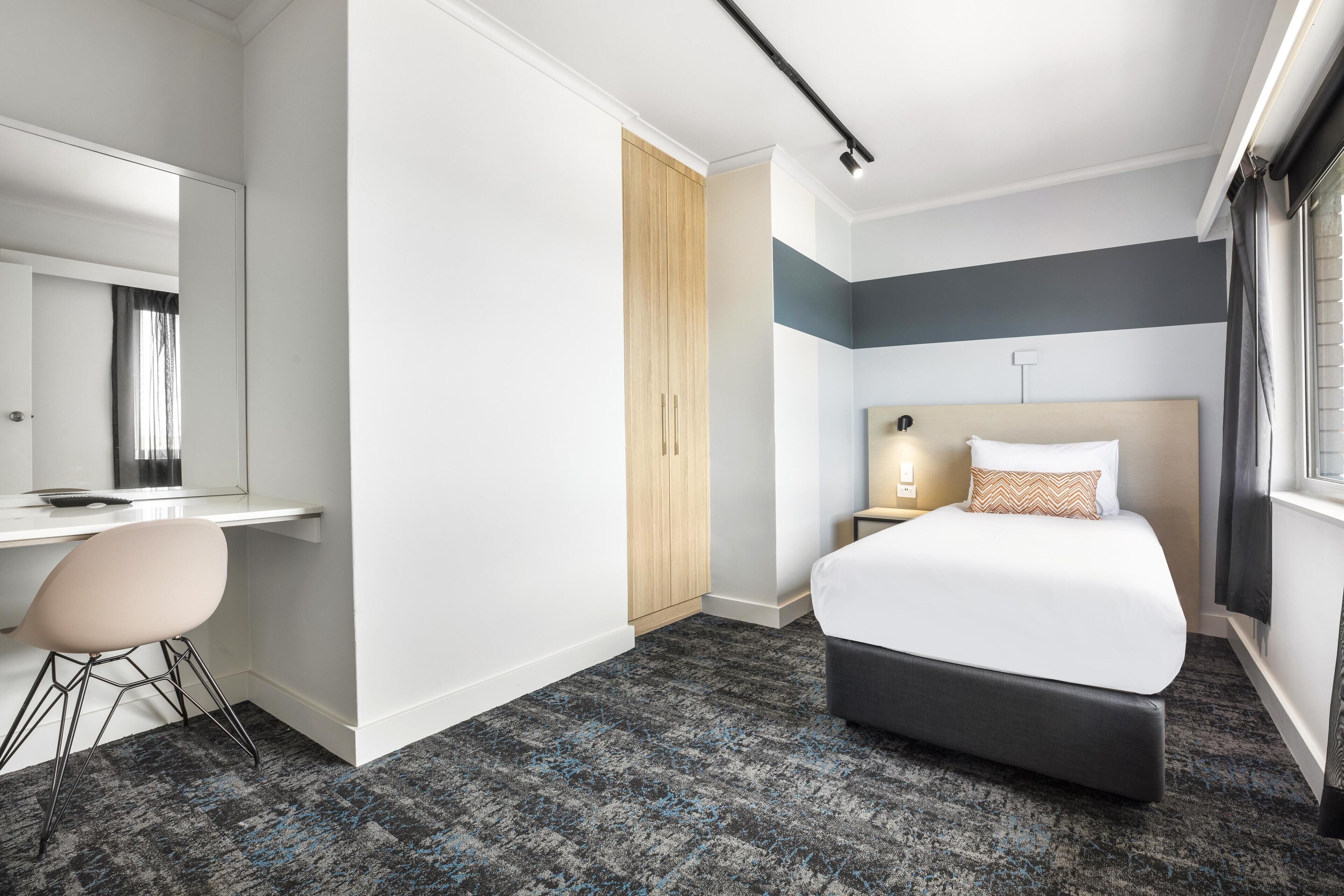 Habitación de hotel con cama, escritorio, silla, armario y ventana.