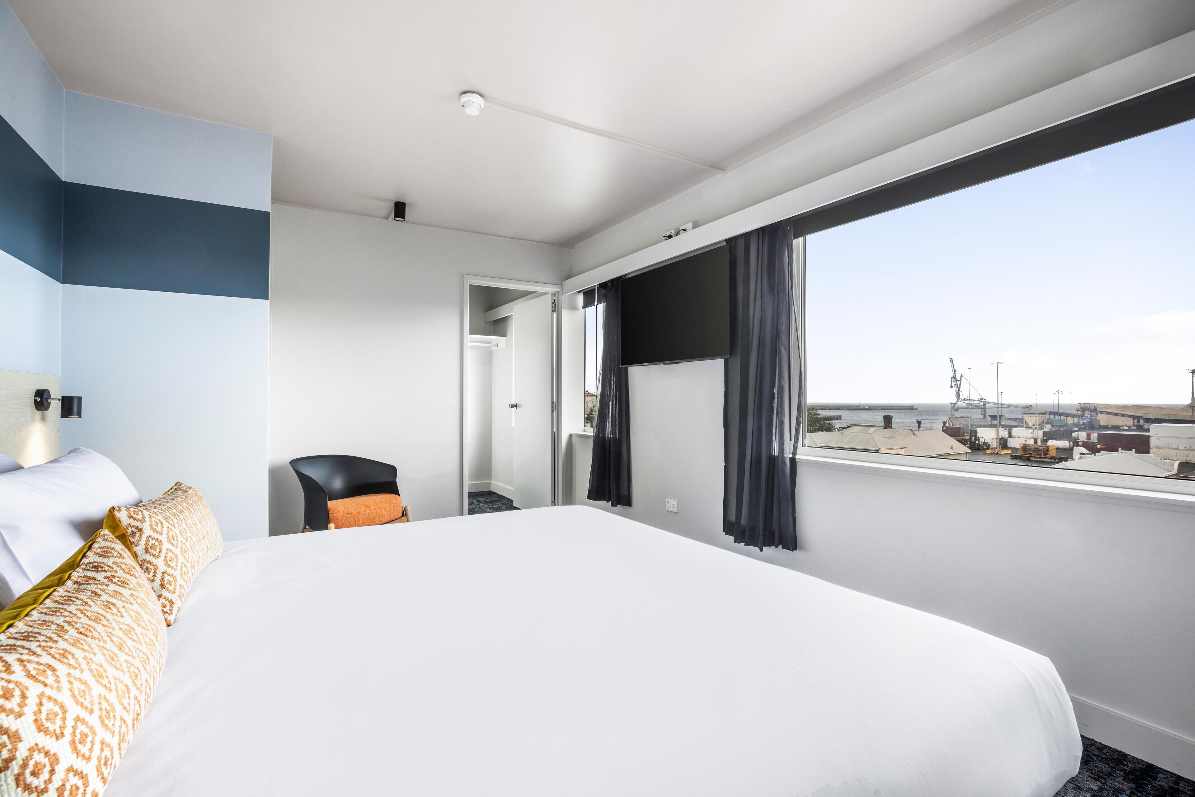 A hotel room with a large bed, a chair, a desk, and a view of the city through a large window.