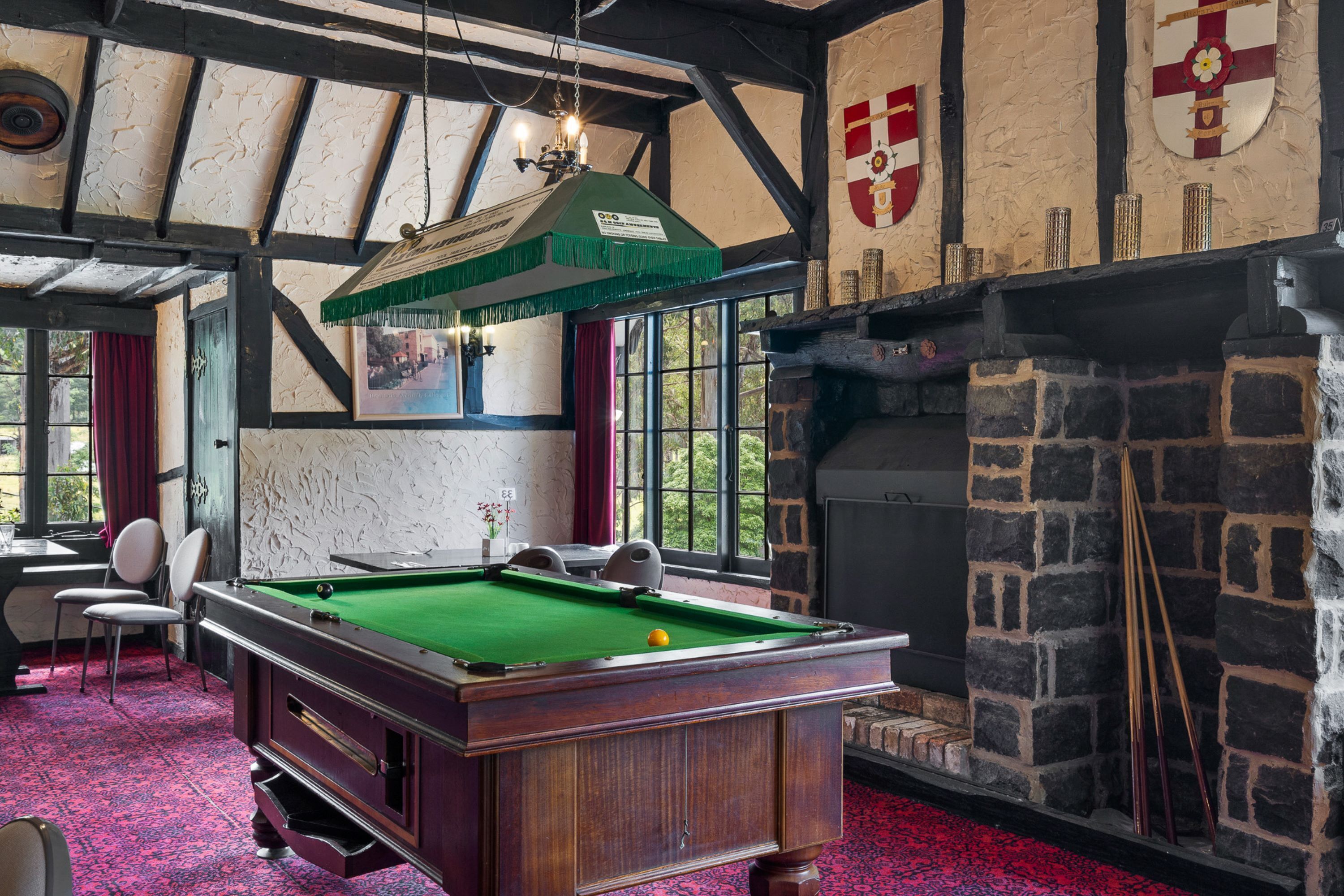 Une table de billard recouverte d’un drap vert, une cheminée avec un manteau en pierre, et une pièce aux poutres apparentes et au tapis rouge.