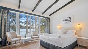 Un dormitorio amplio con una cama grande, dos sillas, una mesita y vistas al exterior a través de ventanas amplias.