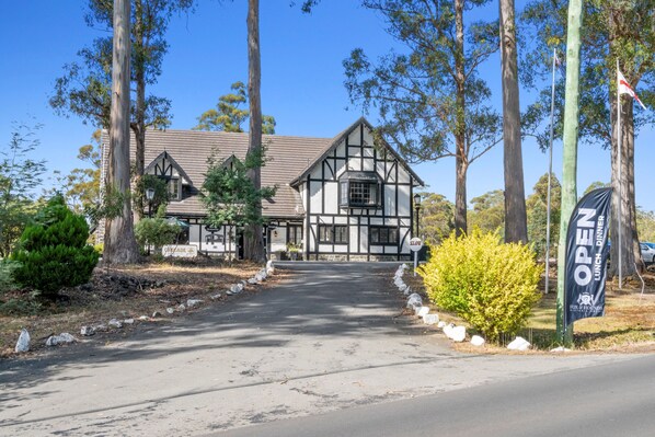 Une maison traditionnelle à ossature bois, avec une allée menant à l’entrée, entourée d’arbres et agrémentée d’un panneau indiquant qu’un événement est en cours.