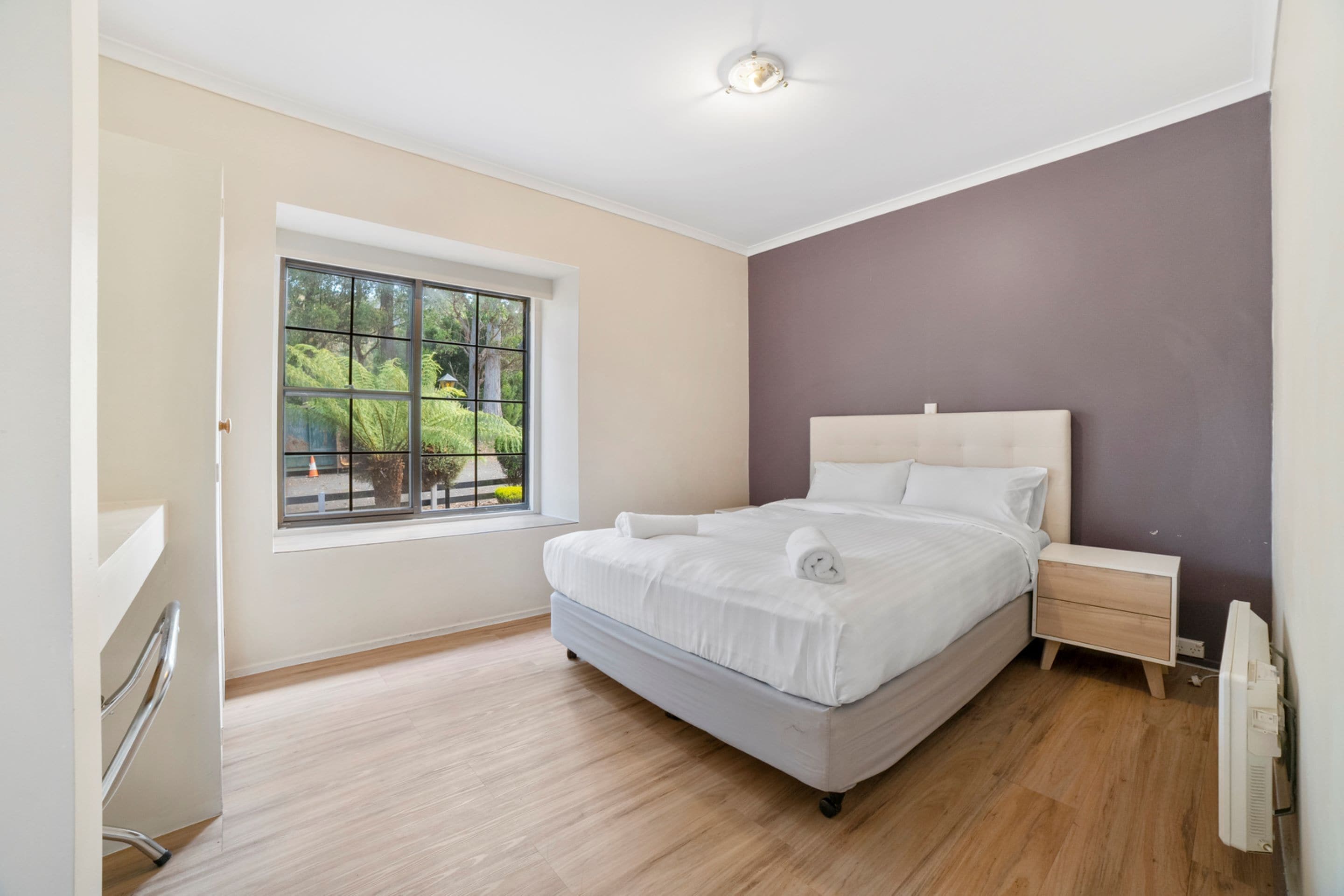 A modern bedroom with a large bed, a bedside table, a window with a view of greenery, and a ceiling light.