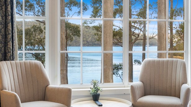 Una habitación con dos sillones beige orientados hacia una ventana grande con vista a un lago. Una mesita redonda con una planta en maceta está en el centro.