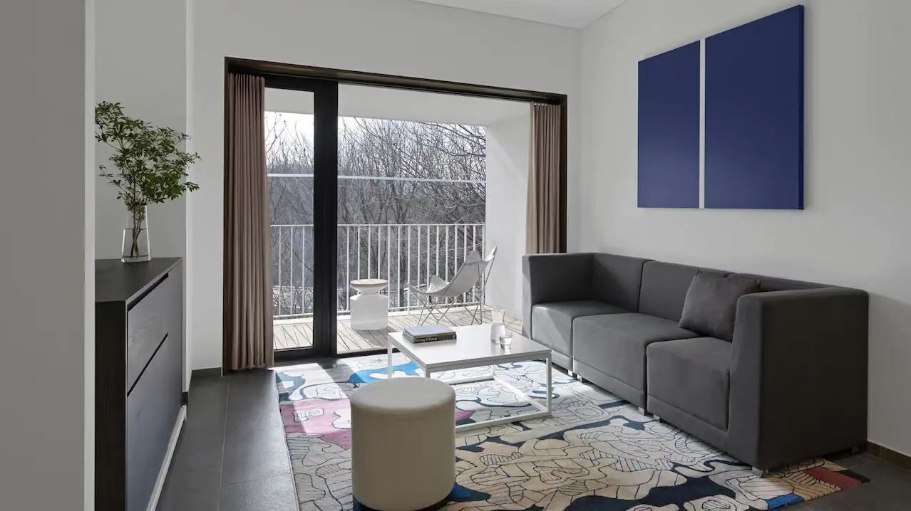 A modern living room with a grey sofa, a small table, and a chair. A large window with curtains leads to a balcony with a view of trees.