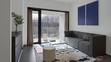 A modern living room with a grey sofa, a small table, and a chair. A large window with curtains leads to a balcony with a view of trees.