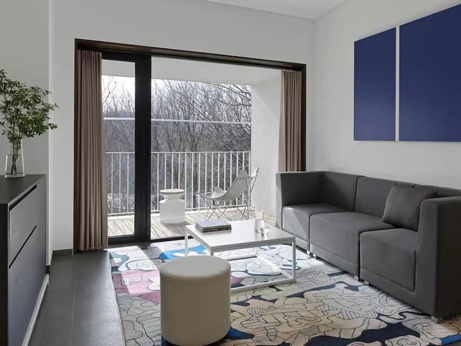 A modern living room with a grey sofa, a small table, and a chair. A large window with curtains leads to a balcony with a view of trees.
