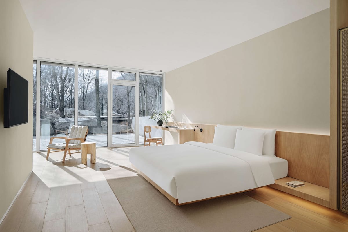 A modern bedroom with a large bed, a desk, and a chair. There is a TV mounted on the wall and a window with a view of trees and rocks.