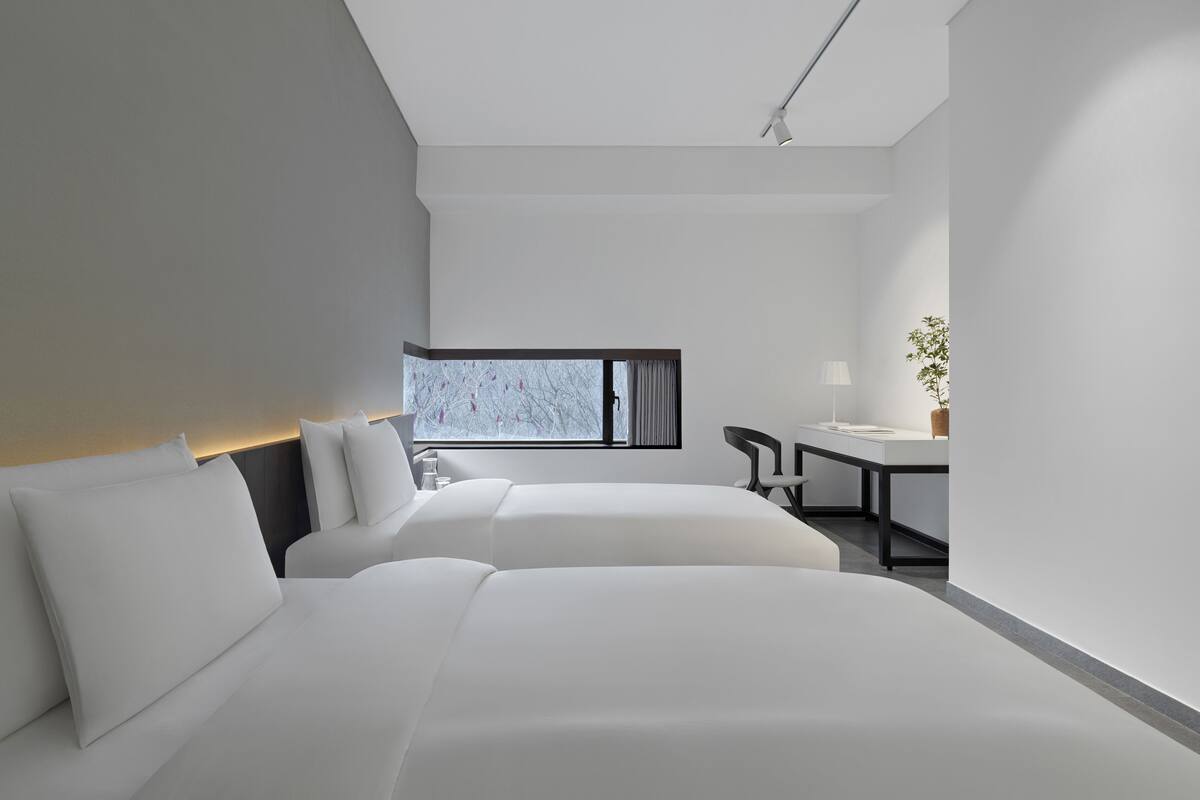 A modern hotel room with two beds, a desk, and a large window.