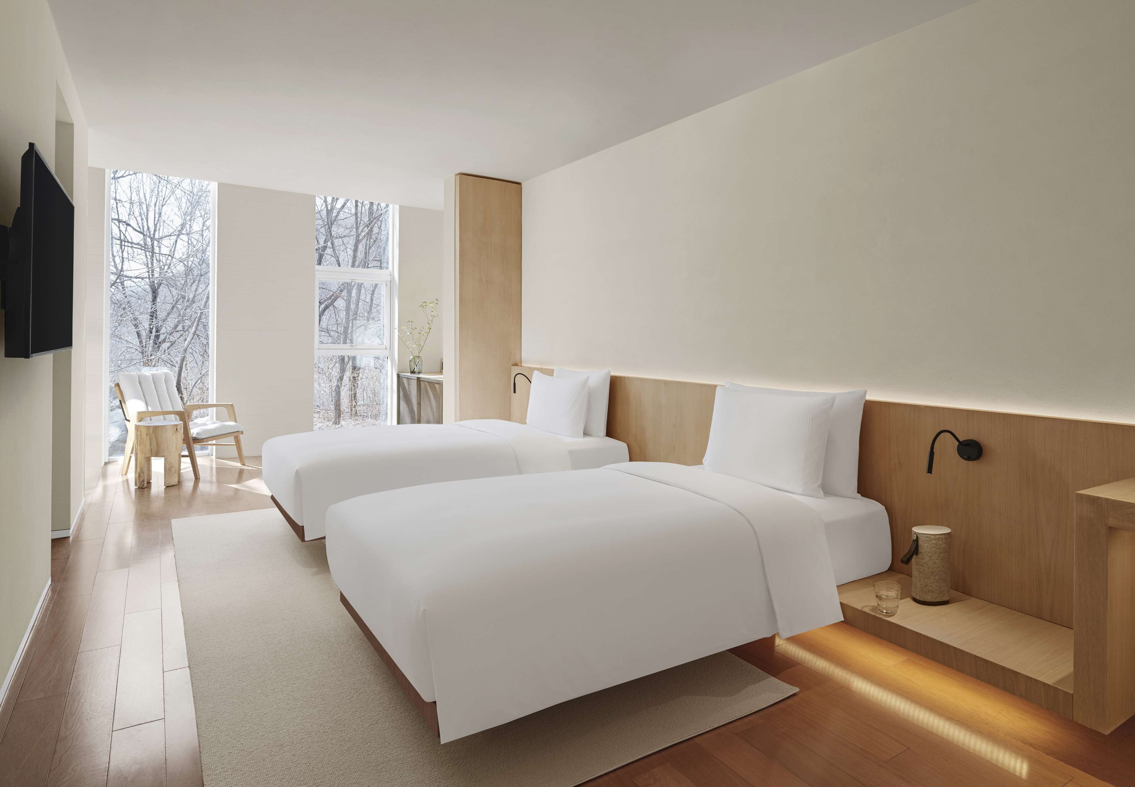 A modern hotel room with two beds, a wooden floor, a large window, and a TV mounted on the wall.