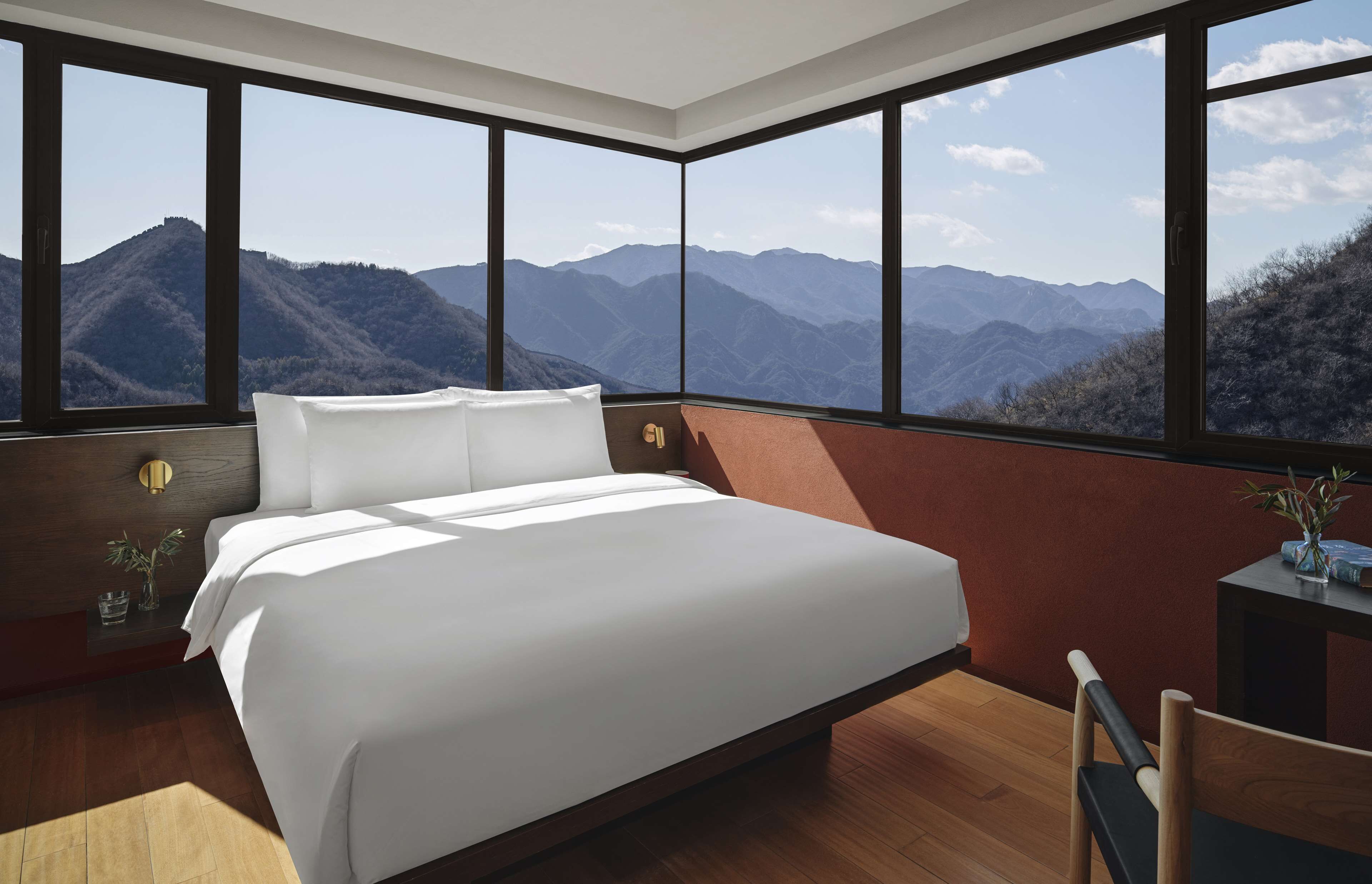A bedroom with a large bed, wooden flooring, a nightstand, a chair, and a view of mountains through large windows.