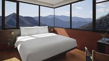 A bedroom with a large bed, wooden flooring, a nightstand, a chair, and a view of mountains through large windows.
