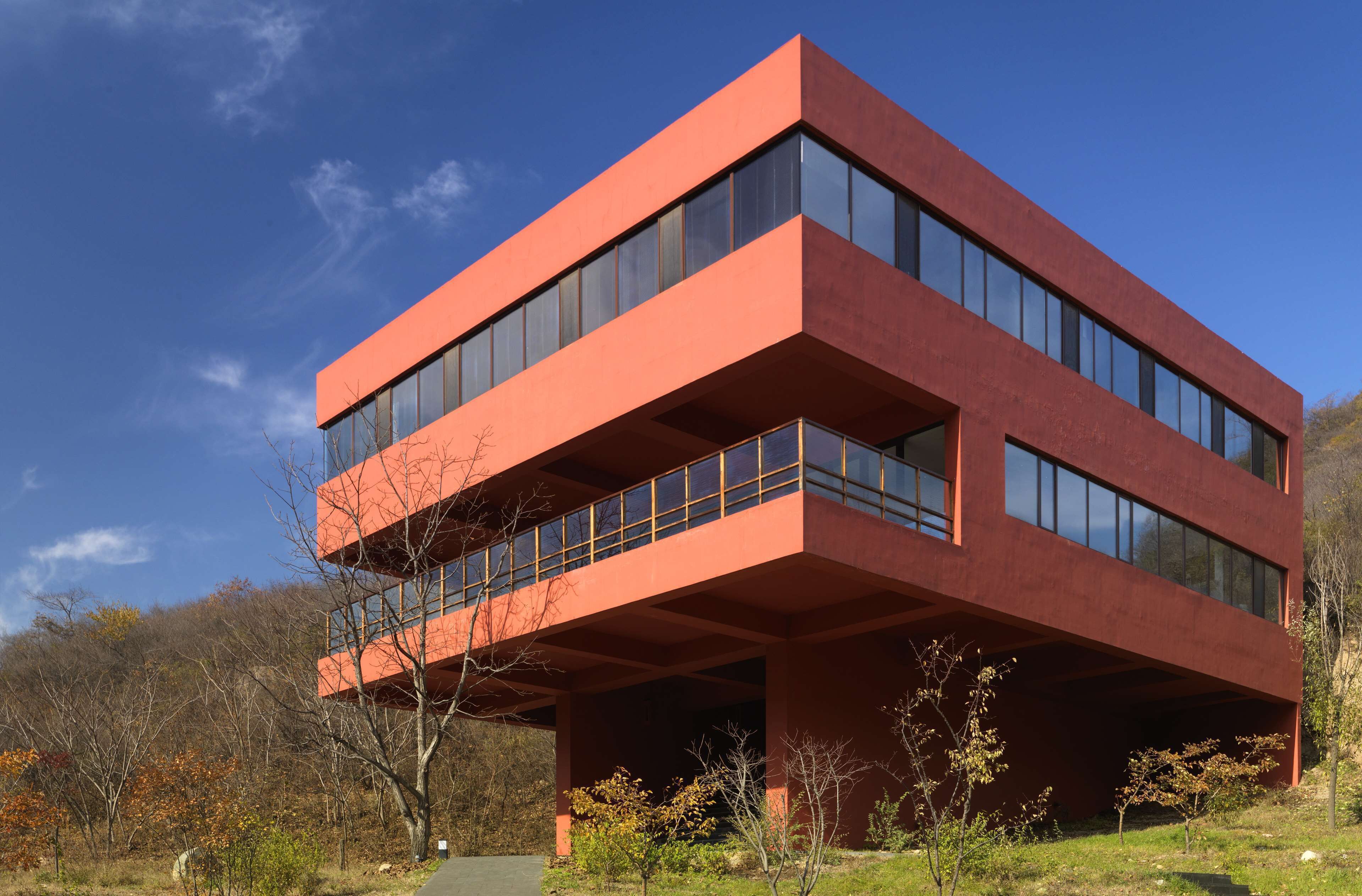Ein modernes, rotes Gebäude mit großen Fenstern und einem Flachdach, umgeben von Bäumen und blauem Himmel.