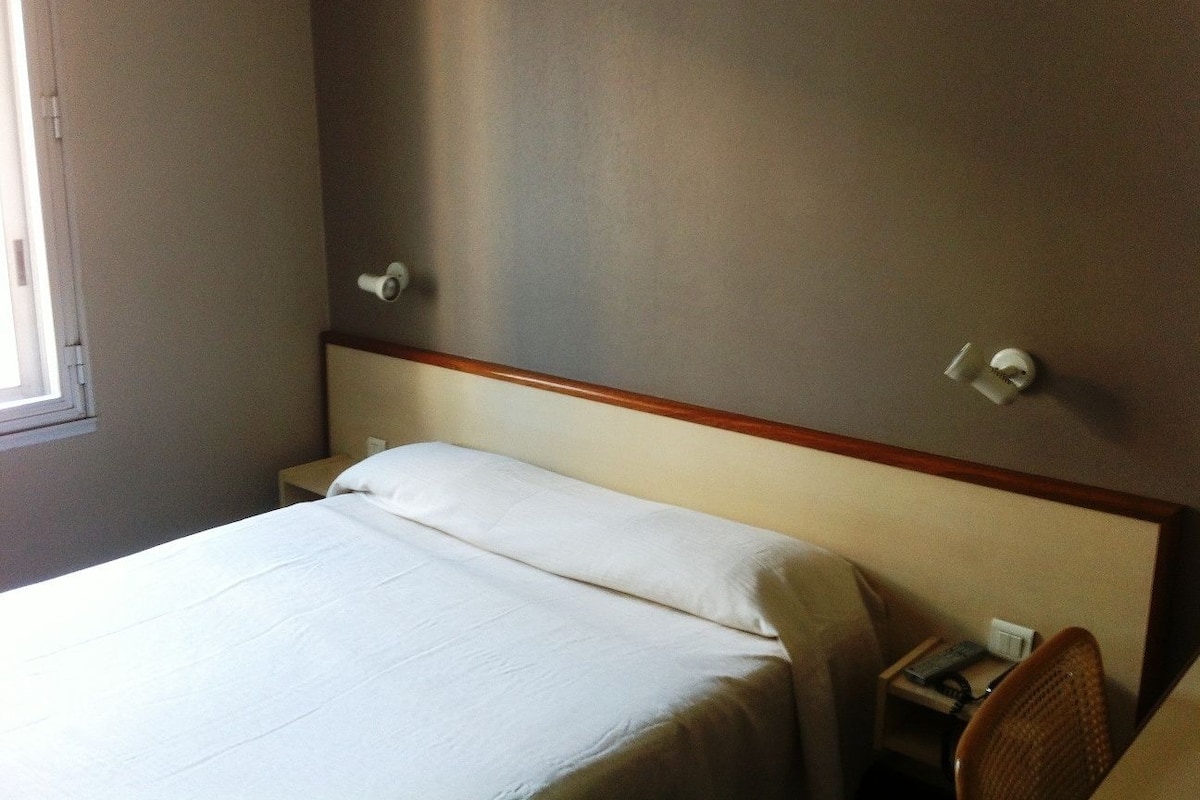 Una habitación de hotel con una cama individual, una mesita de noche, una silla y un ventanal.