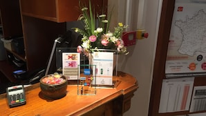 Ein Hotelrezeptionstresen mit Registrierkasse, Blumen in einer Vase, einer Schale mit Süßigkeiten und einer Karte an der Wand.