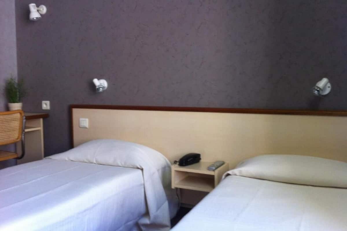 Una habitación de hotel con dos camas individuales, una mesita de noche con teléfono y un estante pequeño.