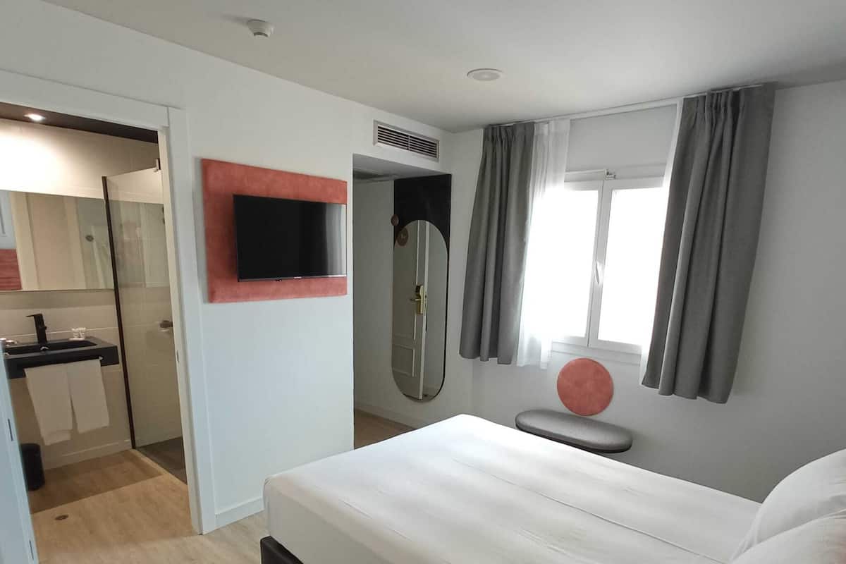 A modern hotel room with a bed, a TV mounted on the wall, a bathroom with a shower, and a window with curtains.