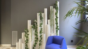 A modern interior with a round marble table, a blue armchair, and a green chair. There are tall white columns with greenery, a projector on the ceiling, and a window with natural light. - Hotel Inca (Zaragoza)