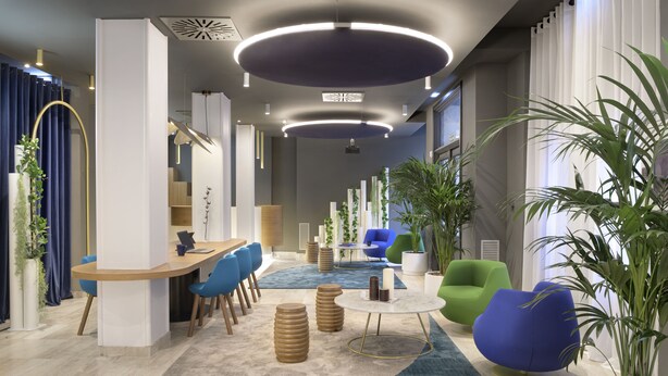 A modern interior with a reception desk, blue and green chairs, a round coffee table, and potted plants. - Hotel Inca (Zaragoza)