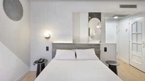 A modern hotel room with a large bed, a bedside telephone, a small table, a round mirror, and a door with frosted glass panels. - Hotel Inca (Zaragoza)