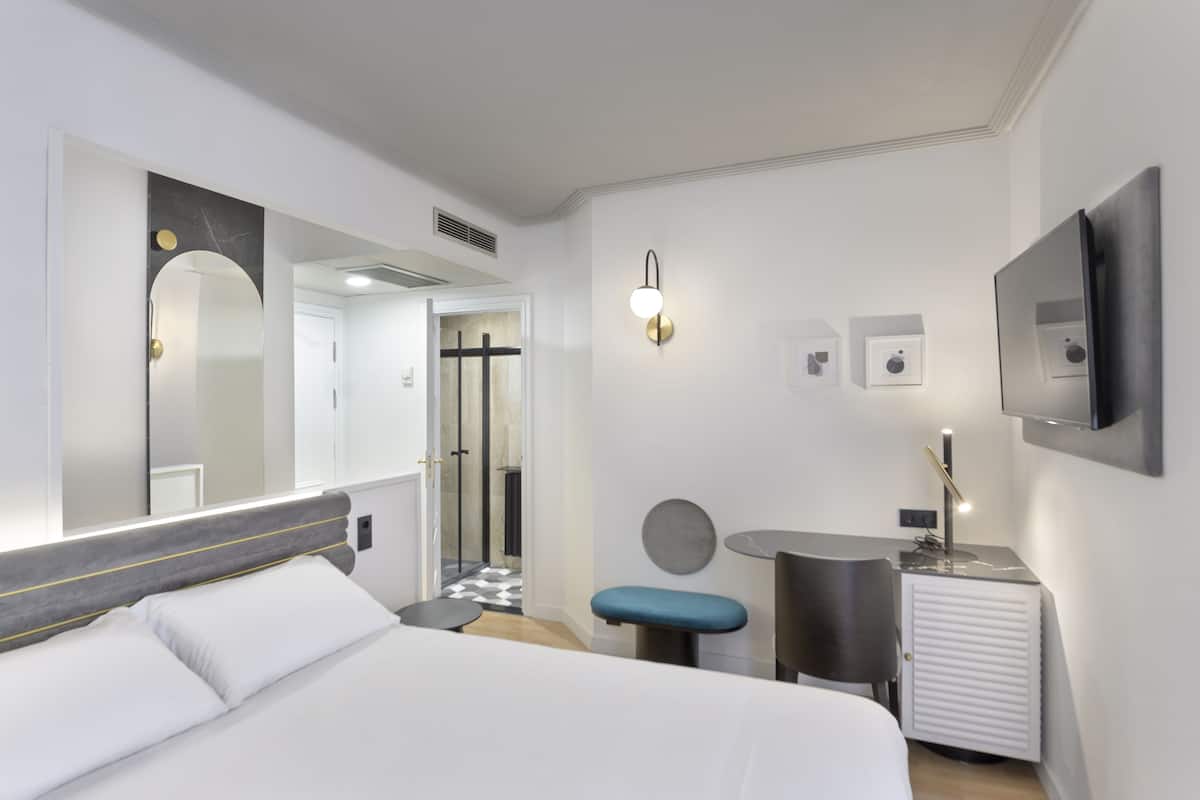 A modern hotel room with a large bed, a desk, a chair, a small table, and a mirror.