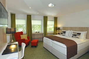 Habitación de hotel con cama, escritorio con lámpara, una silla roja y ventana con cortinas.