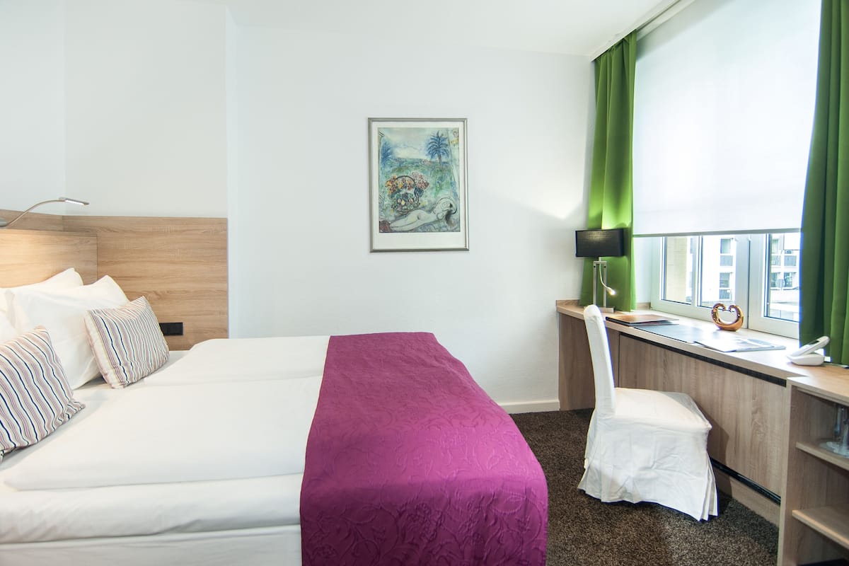 Une chambre d’hôtel avec un lit, un bureau, une chaise, une lampe, un tableau au mur et une fenêtre avec des rideaux.