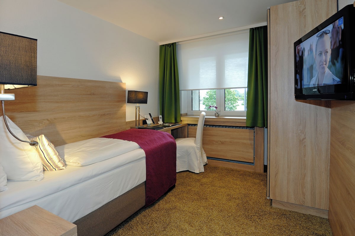 Une chambre d’hôtel avec un lit, un bureau, une chaise, une télévision et une fenêtre avec des rideaux.