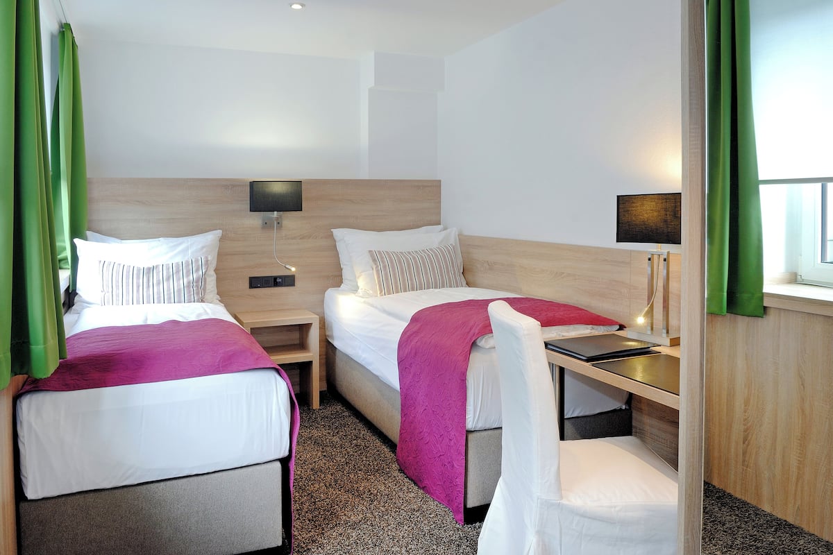 Une chambre d’hôtel avec deux lits, un bureau et une chaise. Il y a deux tables de chevet avec des lampes.