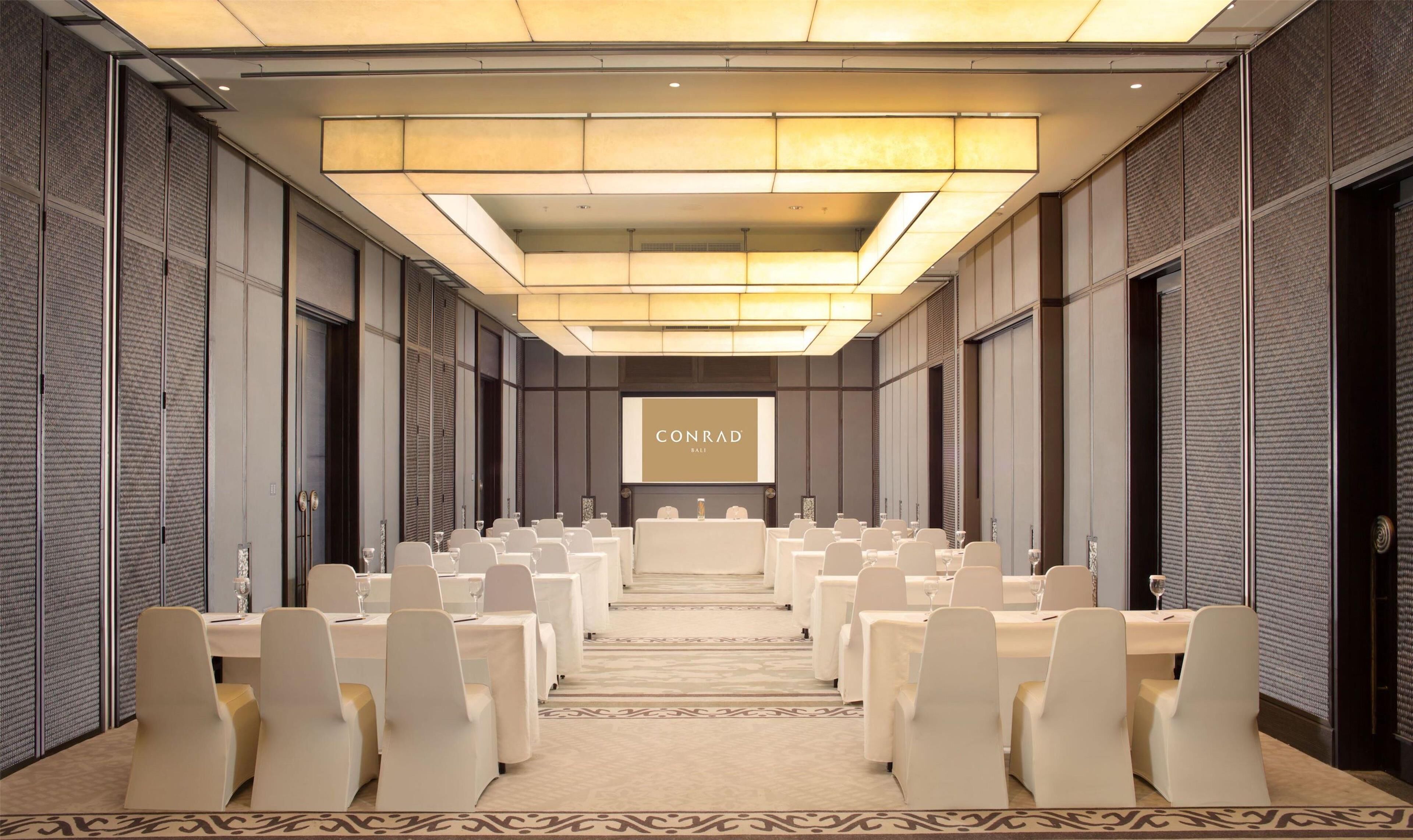 A conference room with rows of white tables and chairs, a large screen displaying 'CONRAD BALI', and a patterned carpet.