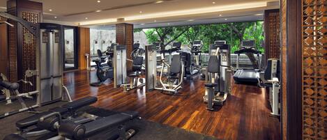 Un gimnasio con diferentes máquinas de ejercicio, suelo de madera y vistas al exterior con vegetación.