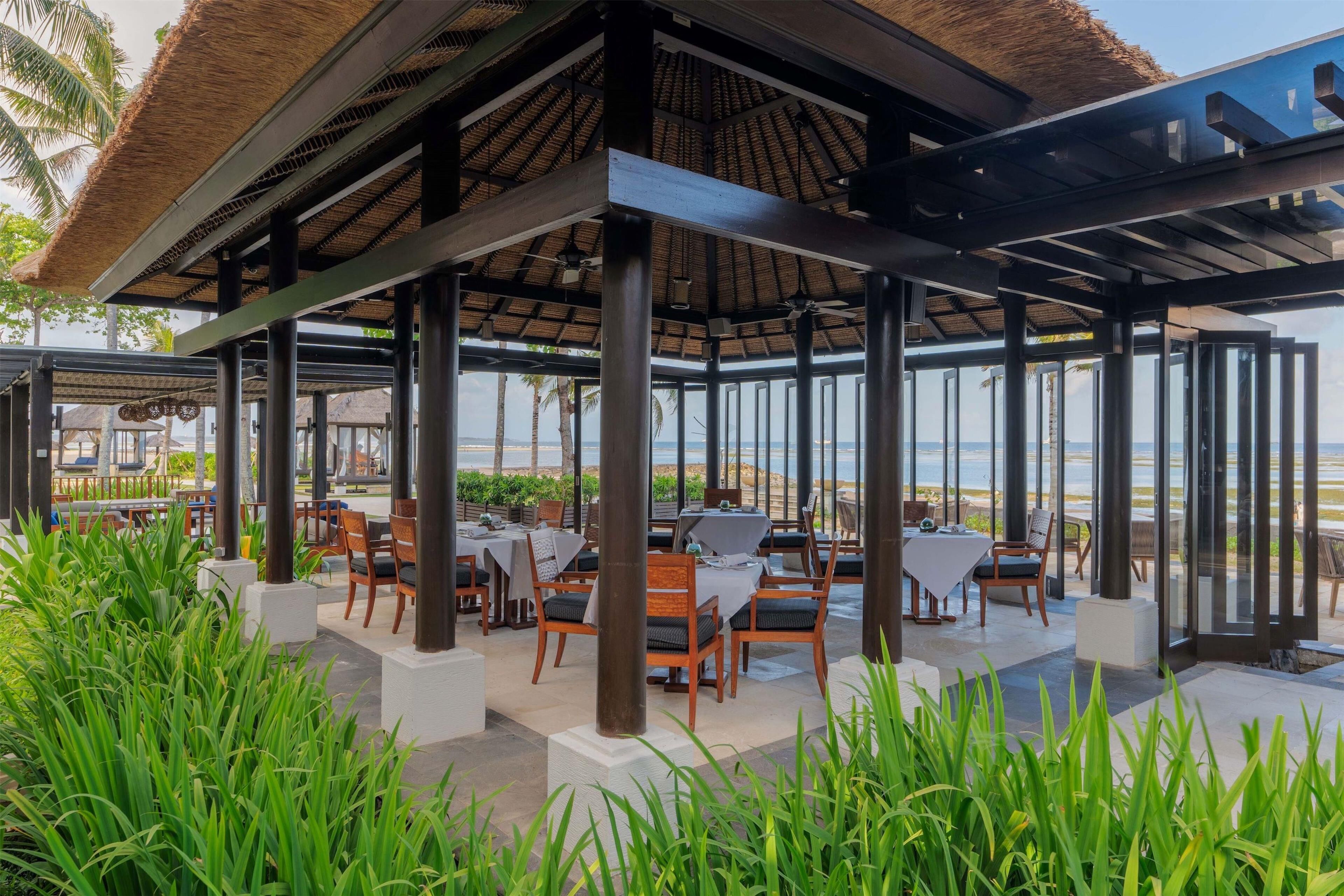 An outdoor dining area with tables and chairs, a thatched roof, and a view of the beach.