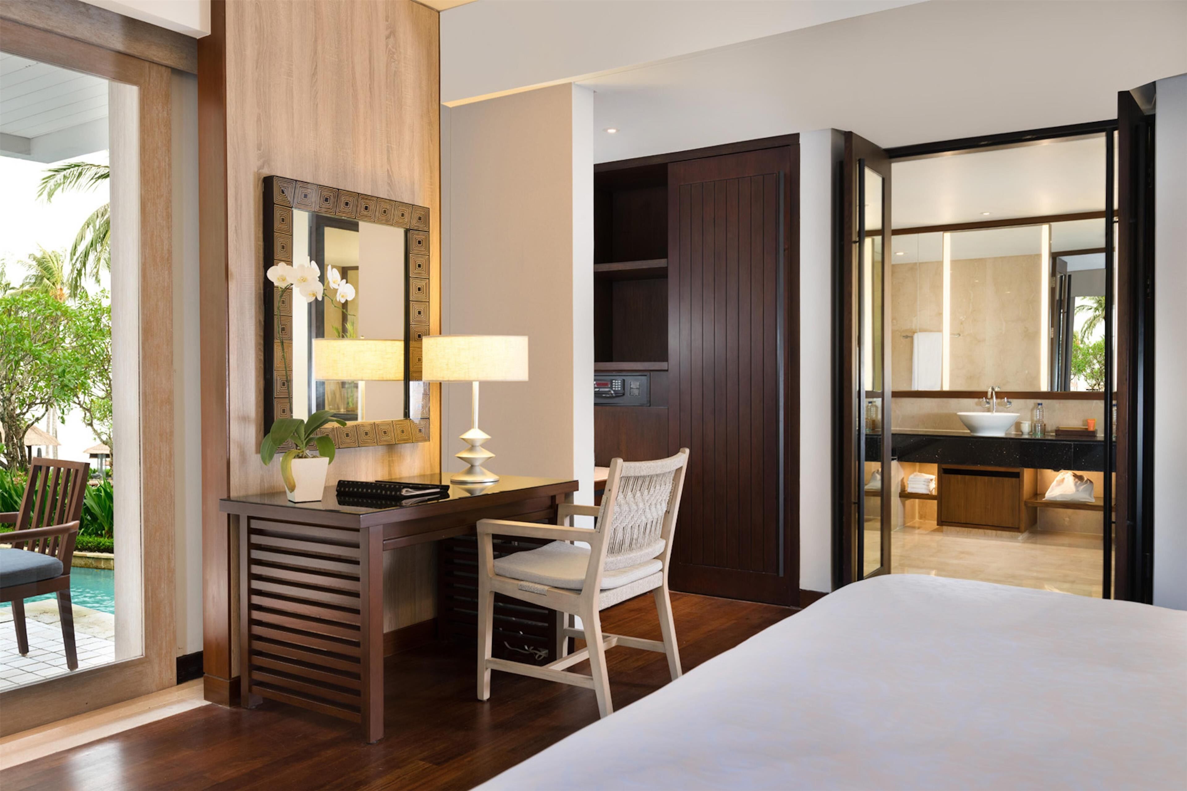 Quarto de hotel com cama, escrivaninha com luminária, cadeira, espelho grande e vista para a piscina e área externa.