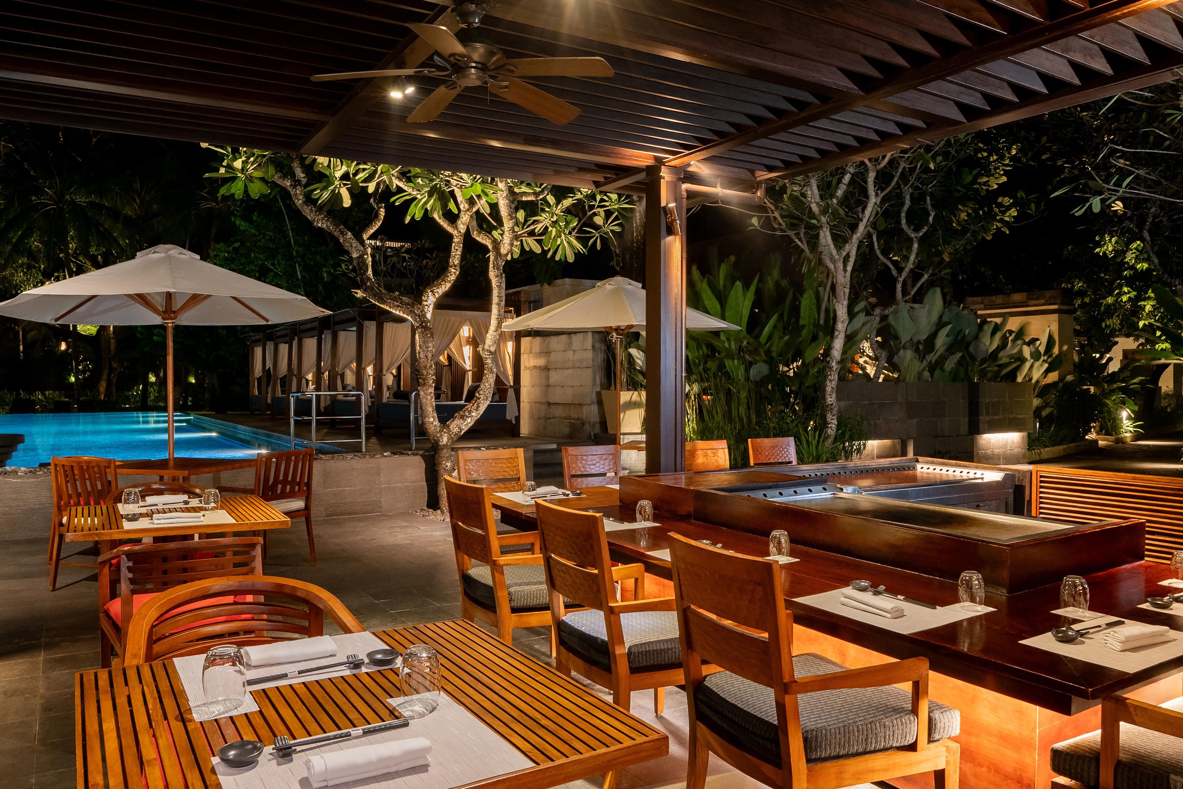Un área de comedor al aire libre con mesas y sillas de madera, sombrillas y una piscina al fondo.