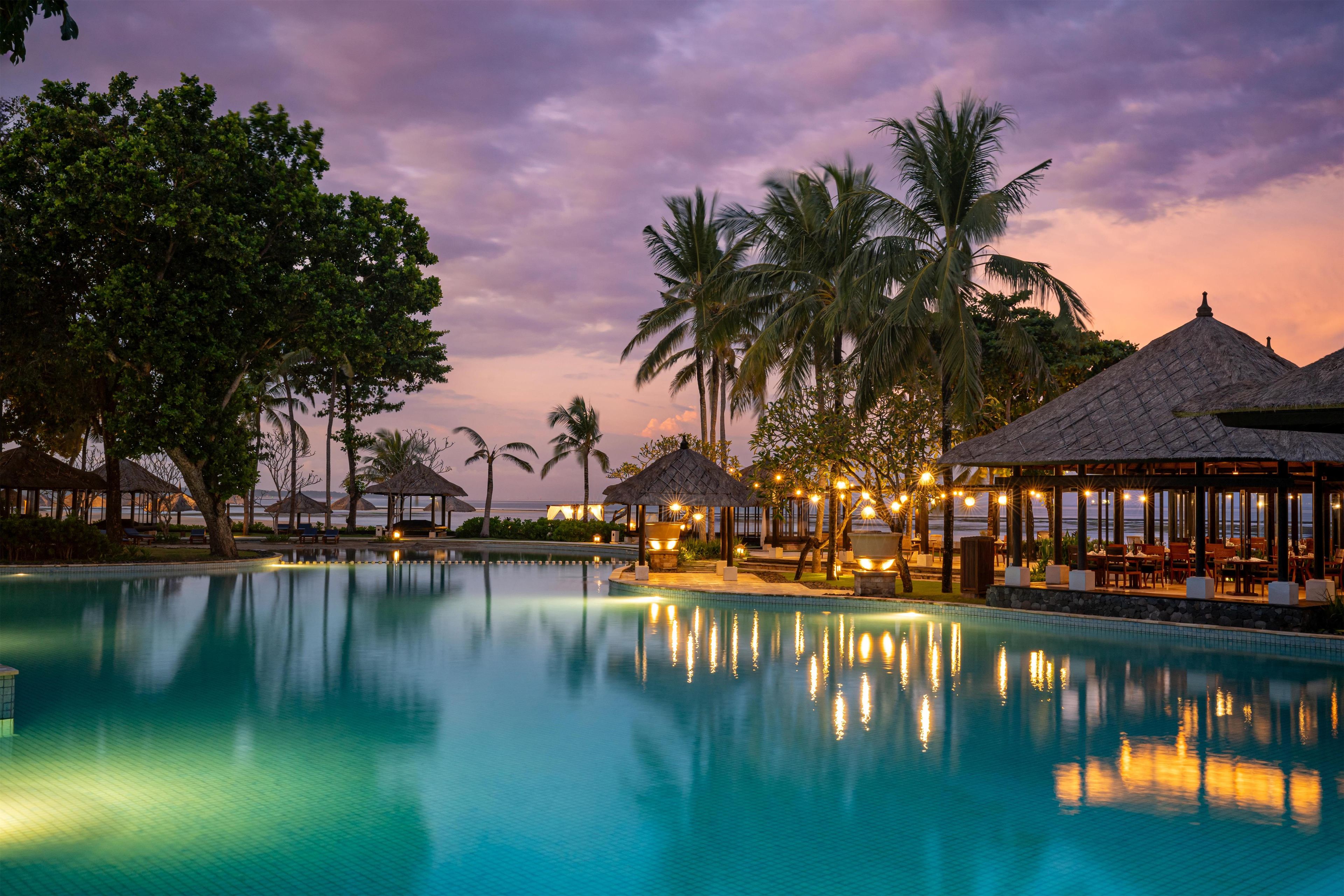 A tranquil pool area with a sunset view, surrounded by palm trees and thatched-roof structures.