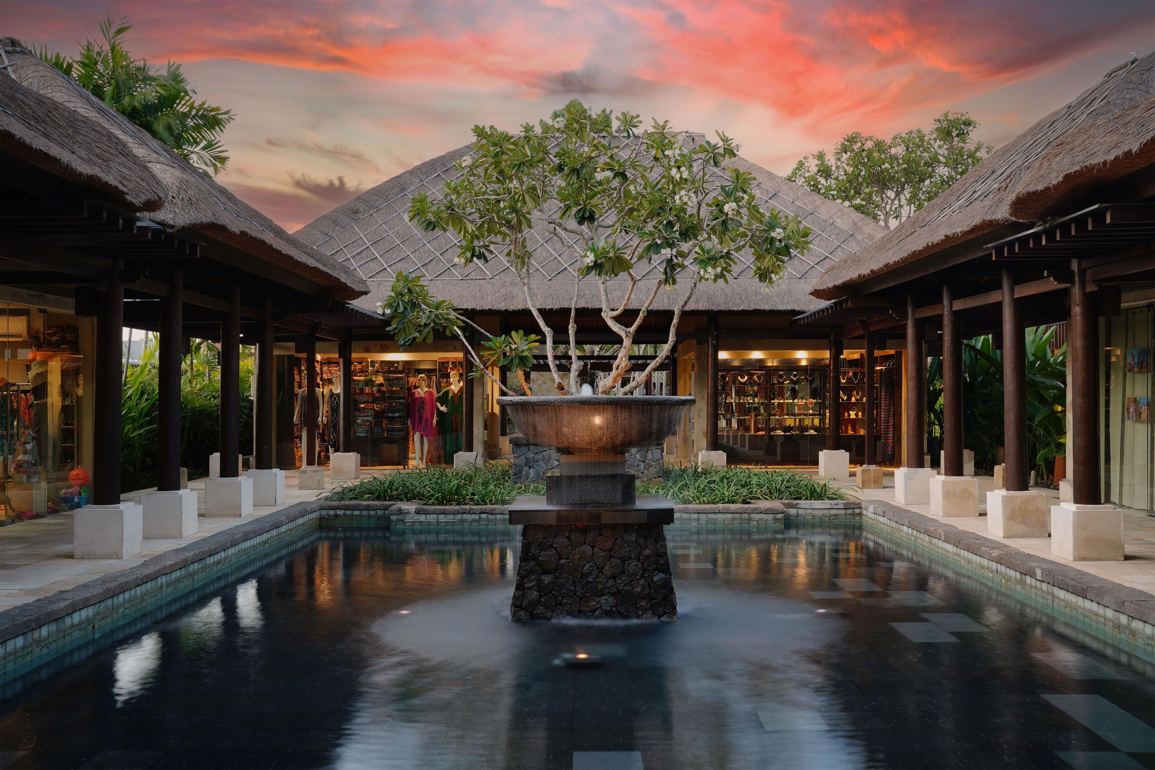A pool with a central fountain, surrounded by thatched-roof structures and a sunset sky.