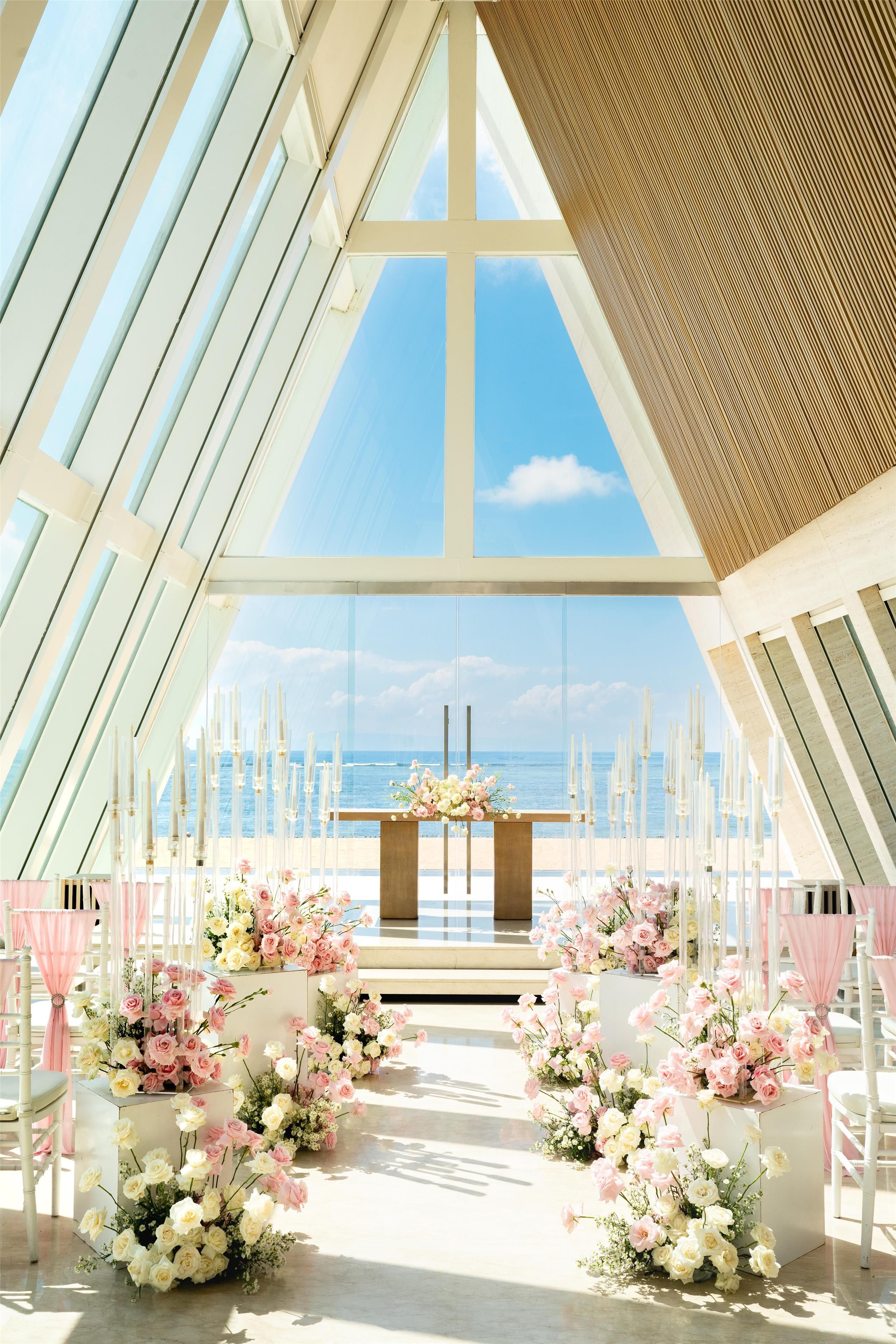 A wedding ceremony setup with a view of the ocean, featuring a central aisle with floral arrangements and chairs on either side.