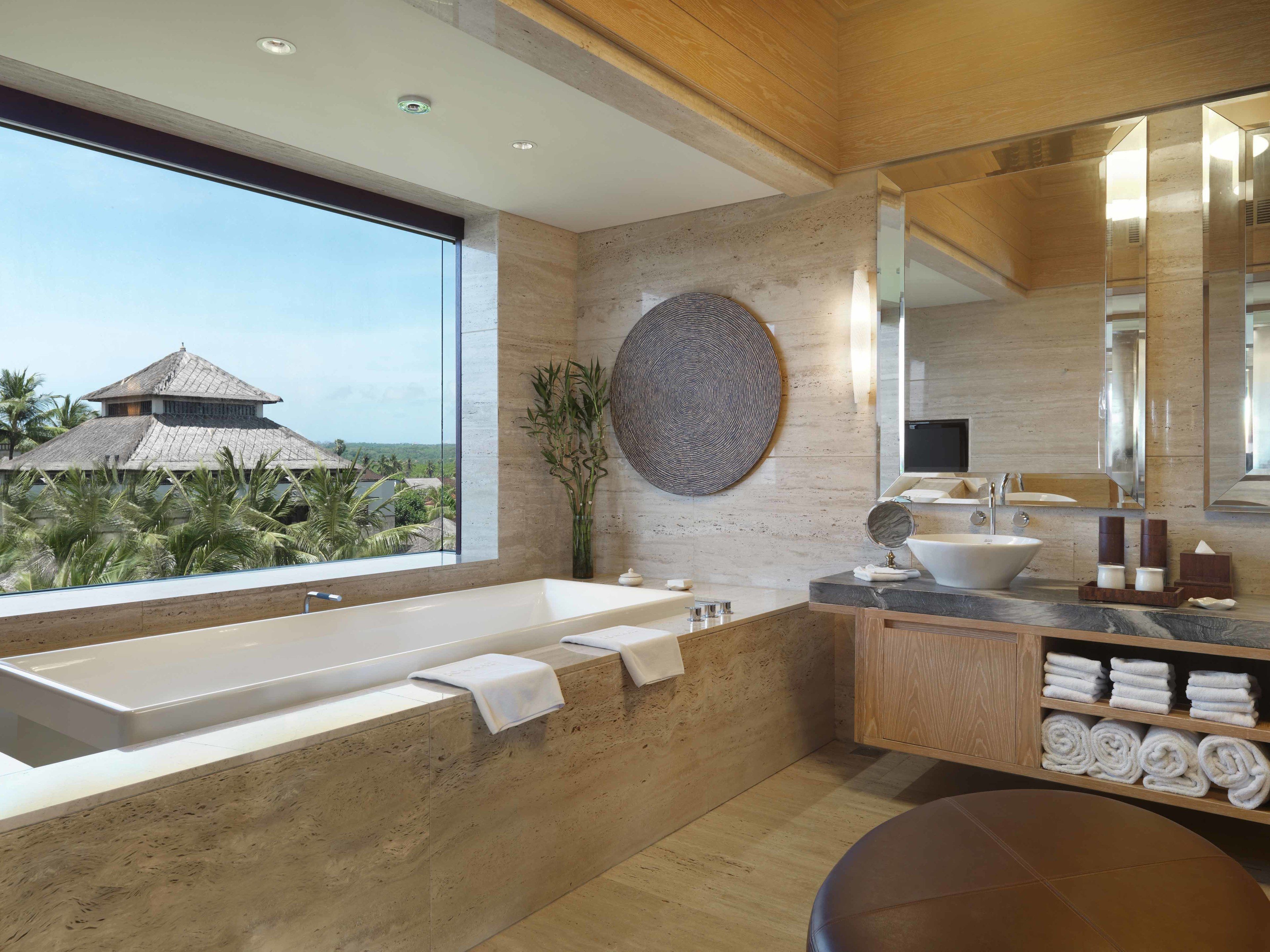 A modern bathroom with a large bathtub, a wooden vanity, and a view of a tropical building.