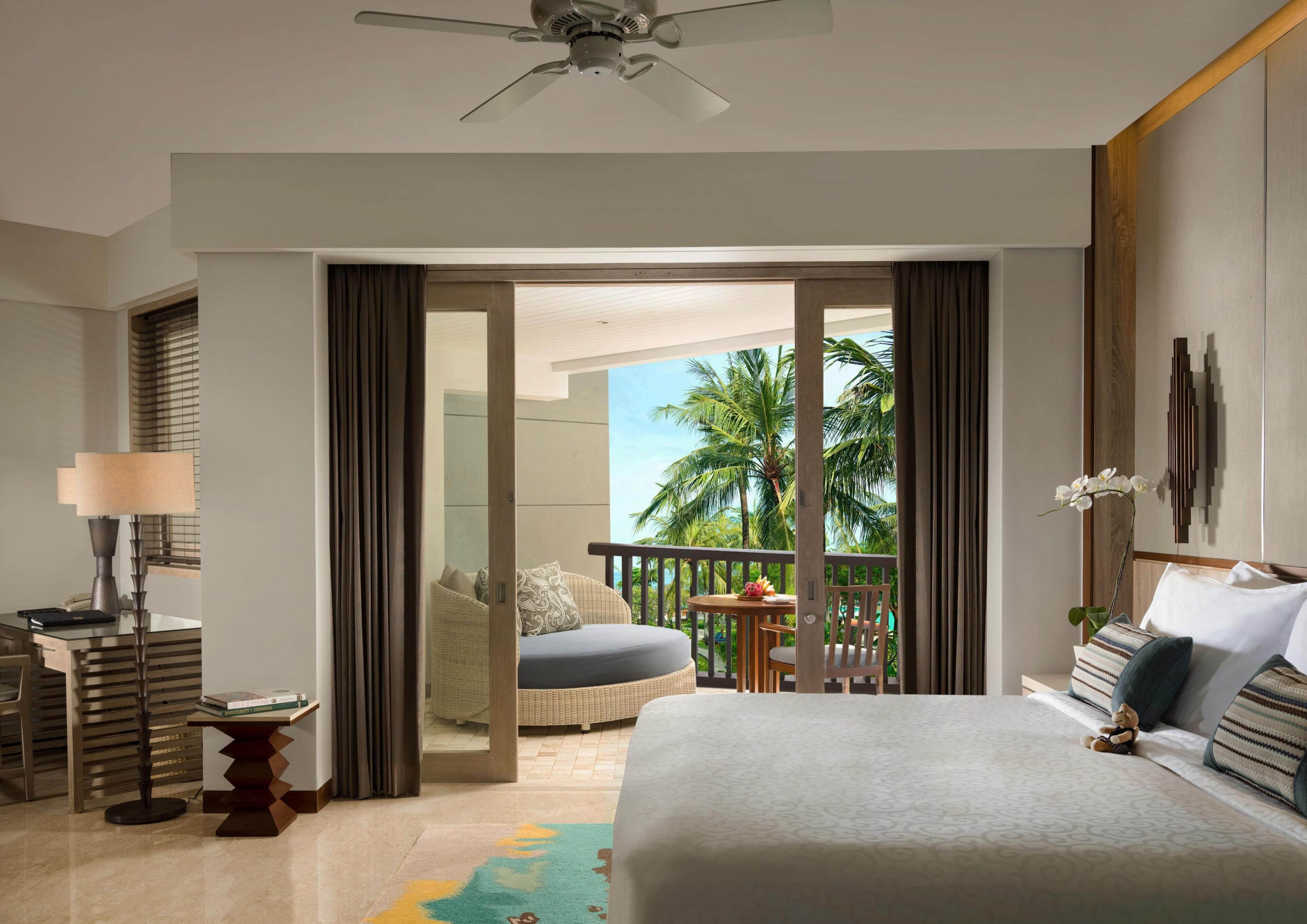 A hotel room with a bed, a desk with a lamp, a chair, a small table with books, a balcony with a view of palm trees, and a ceiling fan.