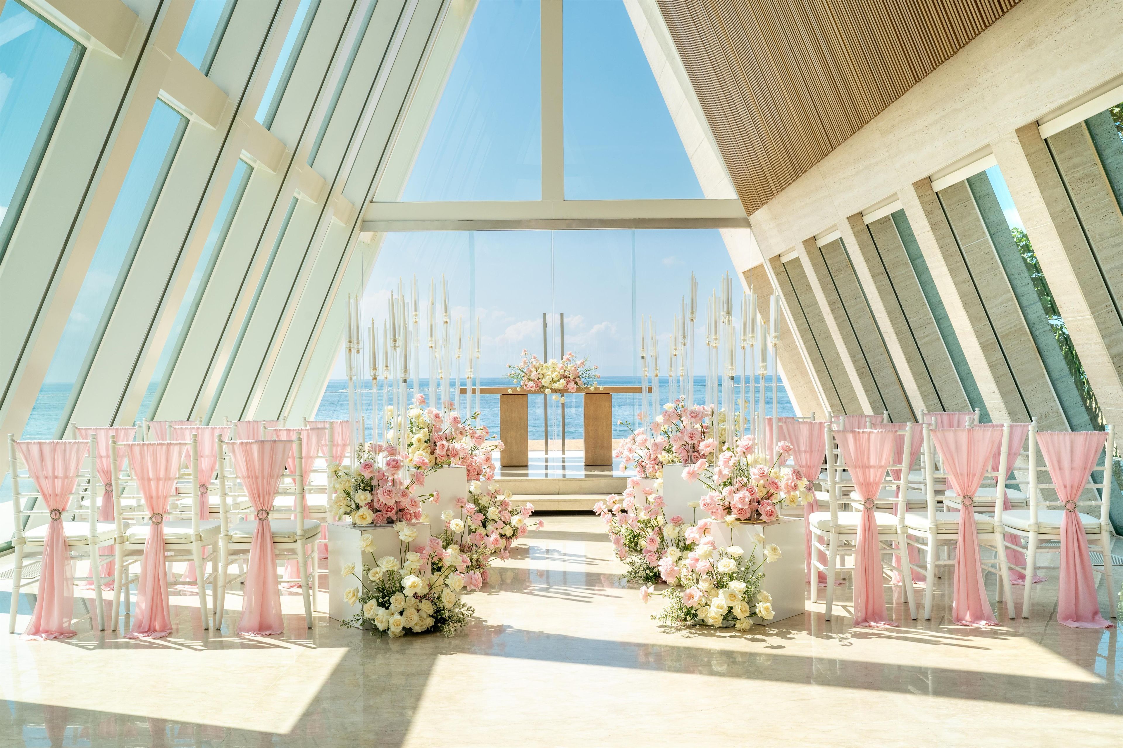 An indoor venue with a clear view of the sea, arranged seating with pink drapes, and floral centerpieces.