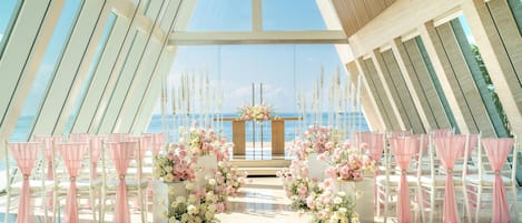 An indoor venue with a clear view of the sea, arranged seating with pink drapes, and floral centerpieces.