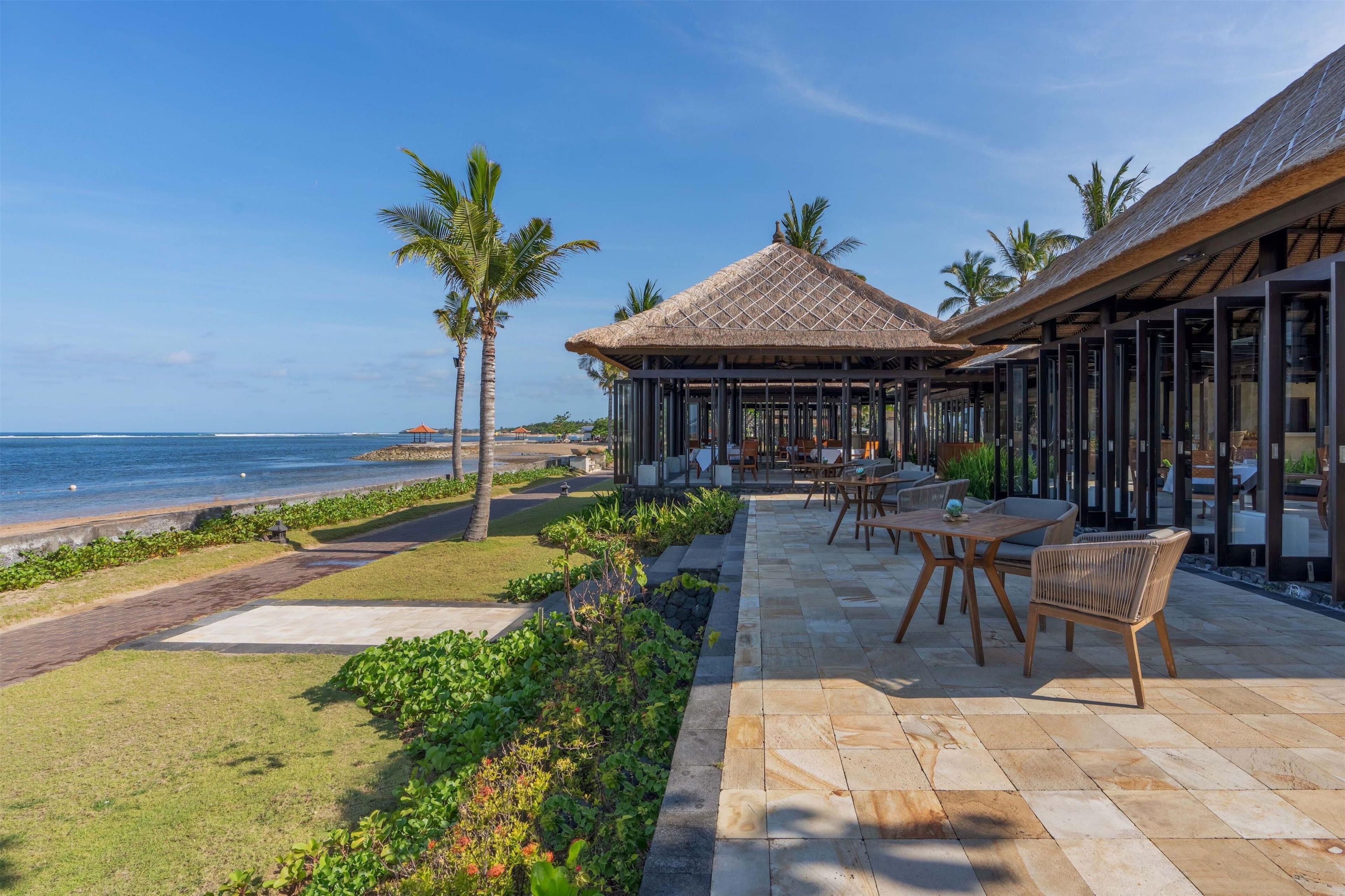 A beachfront patio with a thatched-roof gazebo, outdoor seating, and a view of the ocean.